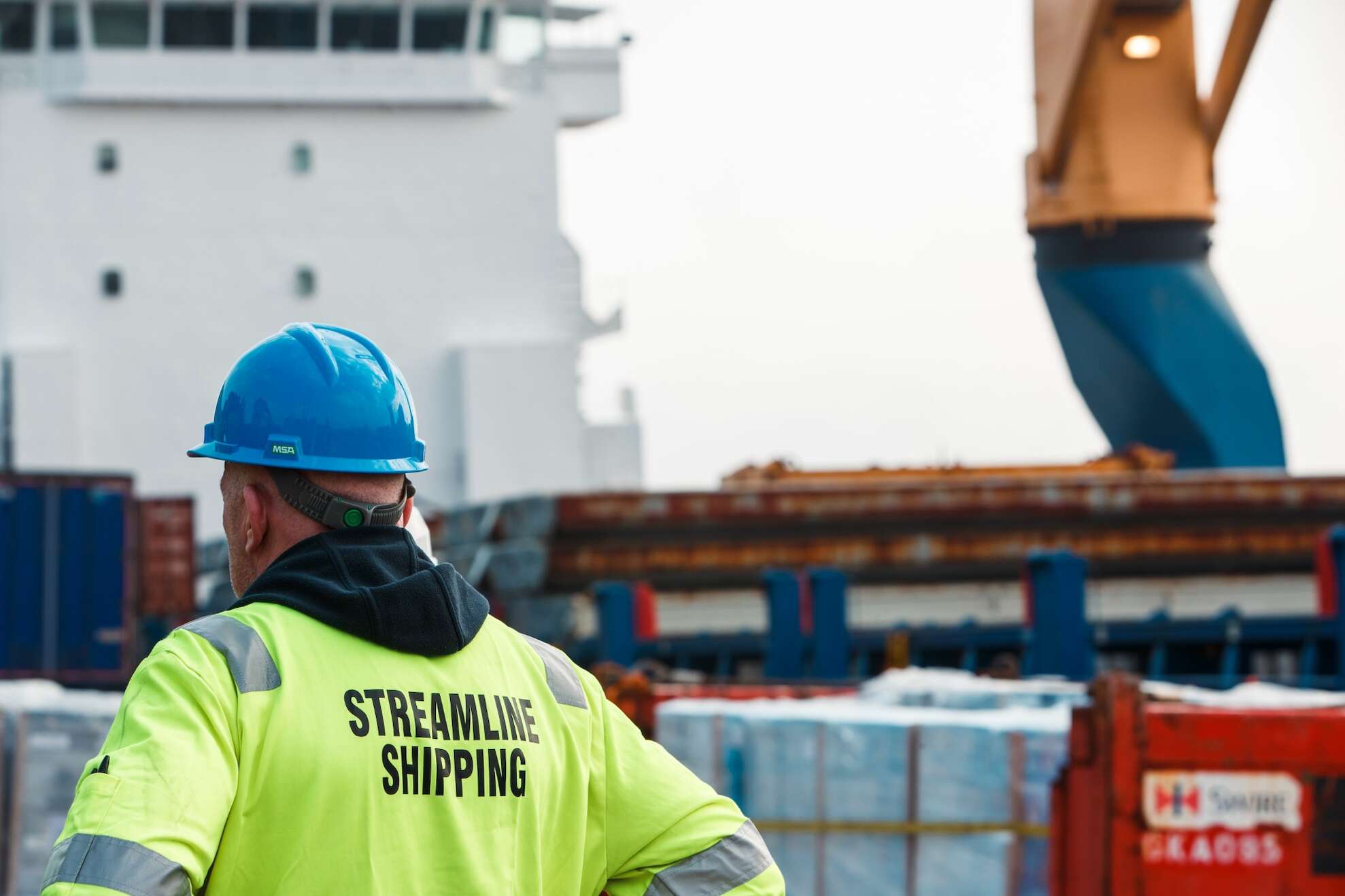 Streamline shipping worker standing, wearing a blue hard hat and a hi vis jacket in front of a ship