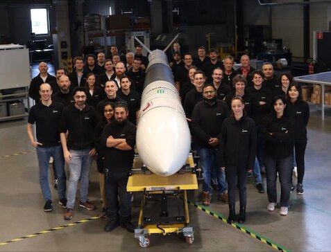 A large group of workers wearing navy blue standing next to a white rocket, smiling to camera