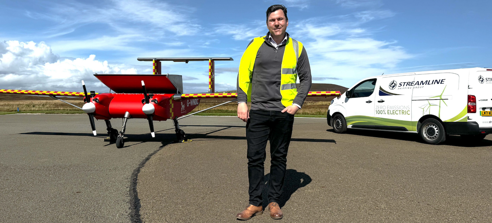 A worker wearing a yellow vest standing outdoors between a white Streamline van and a red drone