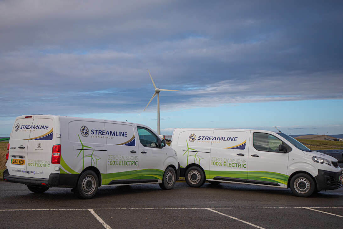 Two white Streamline vans parked on a road with a cloudy sky and wind turbine in the background